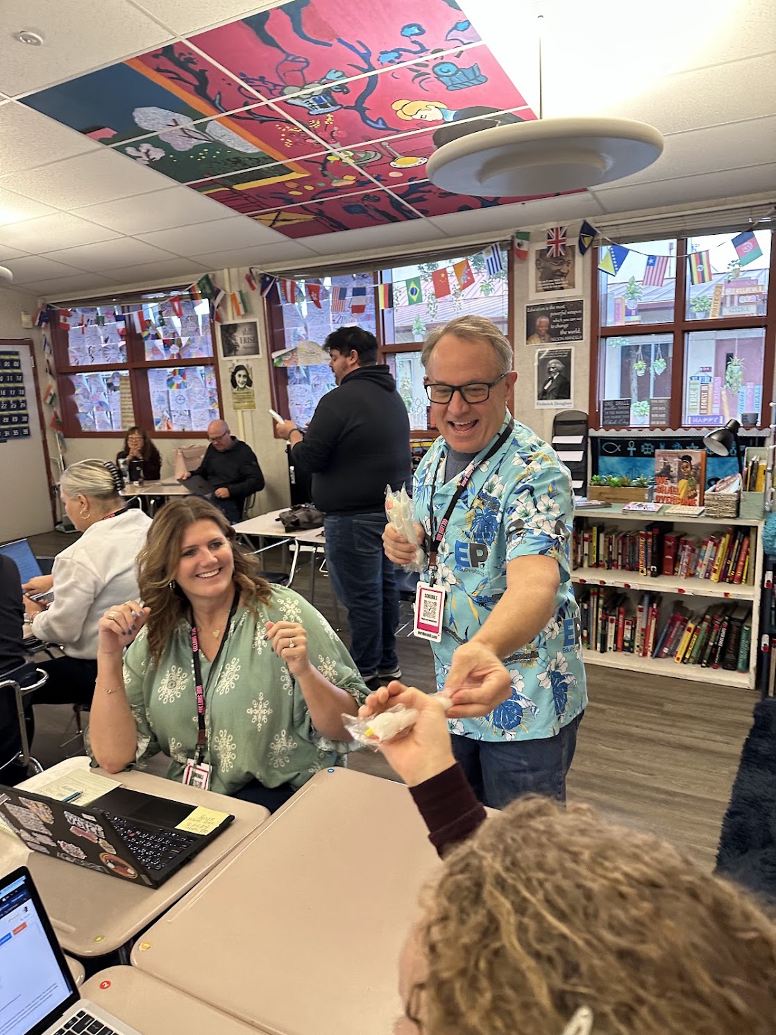 Jon Corippo handing out snacks to smiling Vibe Shift 2025 attendees in a colorful classroom strung with world flags and lined with bookshelves