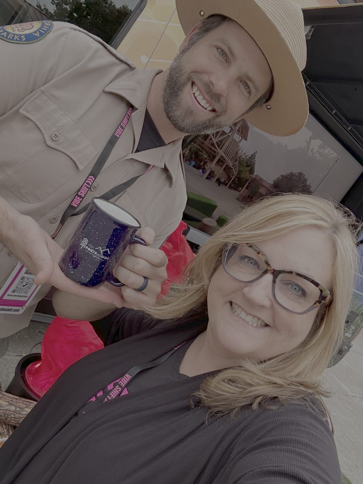 A Vibe Shift 2025 attendee posing outside the venue with a state park ranger, both holding a speckled Vibe Shift enamel camp mug