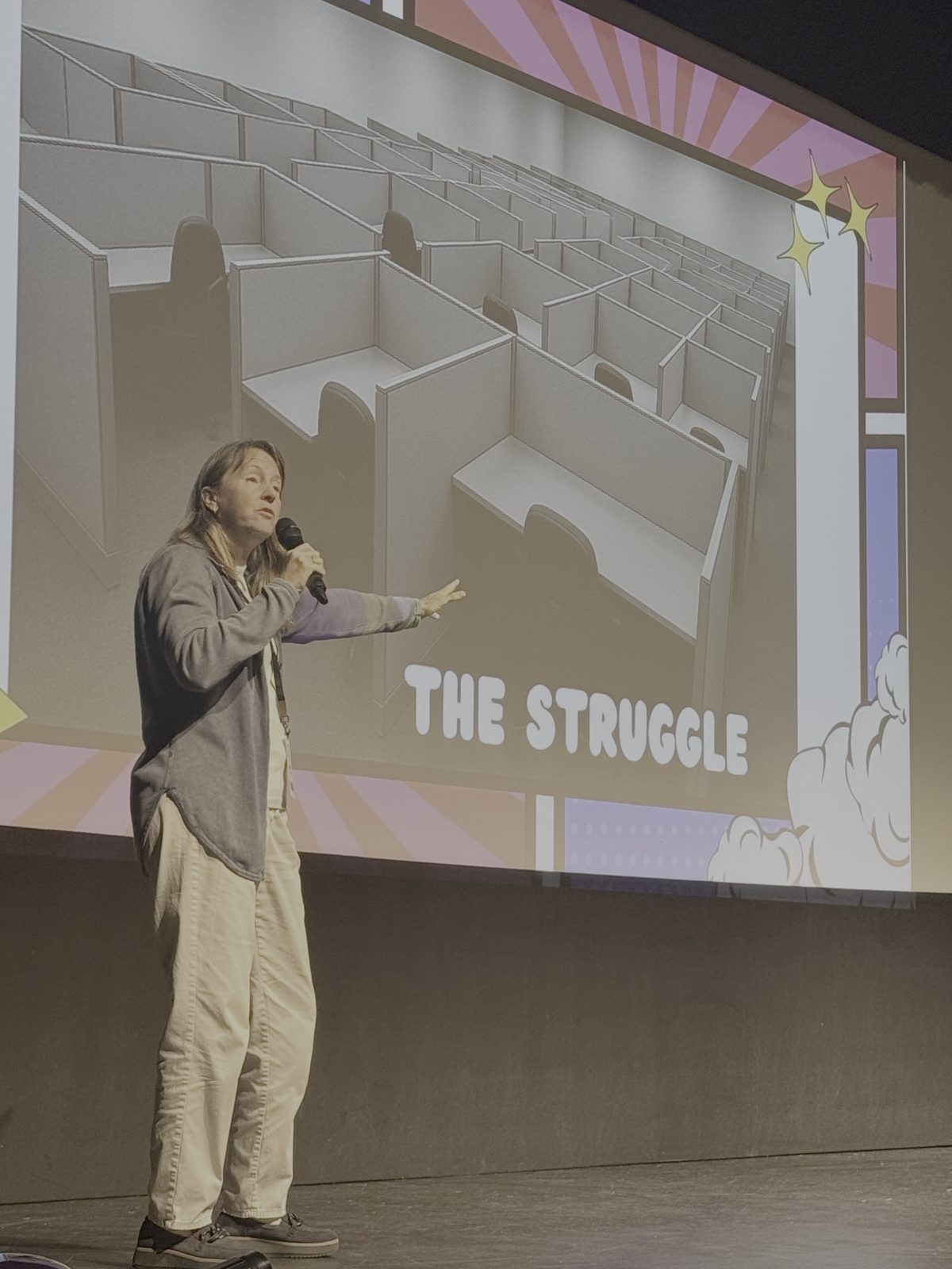 A Vibe Shift 2025 speaker addressing the audience in front of a projected slide titled "The Struggle" showing an empty office cubicle maze