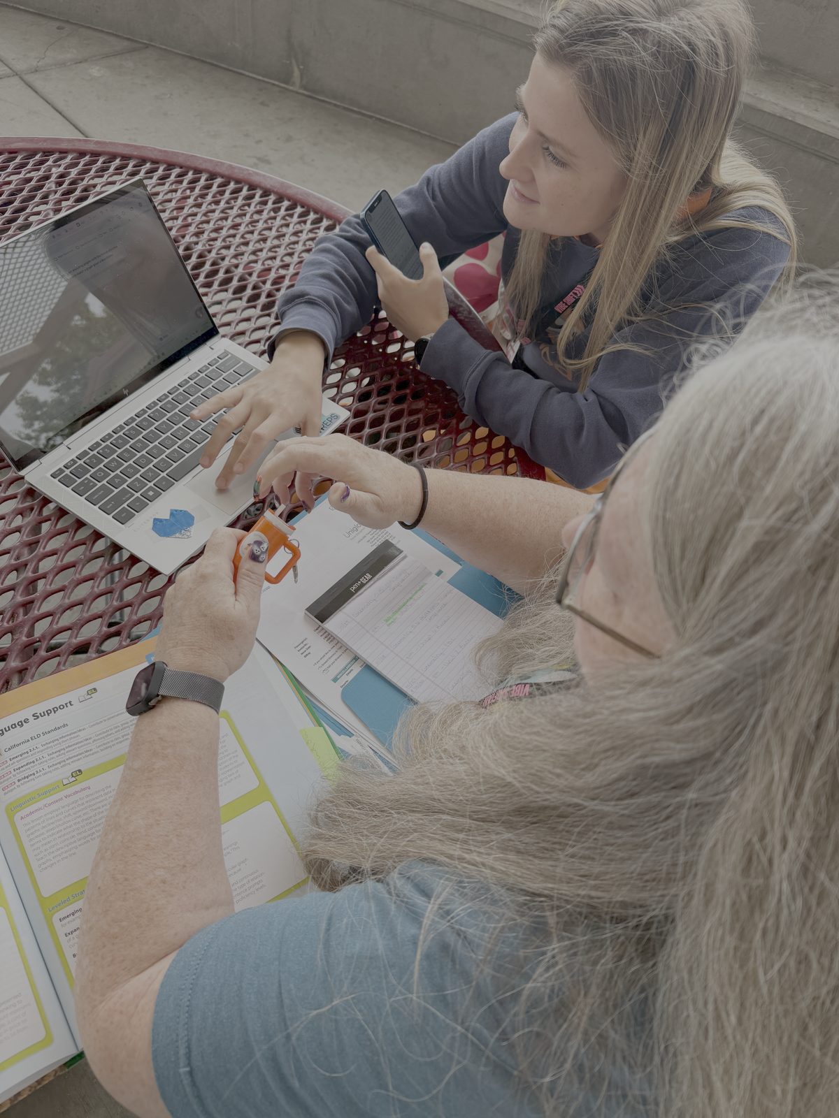 Two Vibe Shift 2025 attendees collaborating outside at a red mesh table with laptops, a phone, and a printed EduProtocols lesson workbook