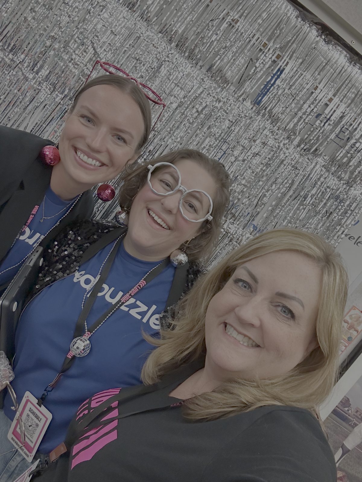 Three Vibe Shift 2025 attendees in matching blue Puzzle.com shirts and disco-ball earrings taking a group selfie in front of a silver tinsel backdrop